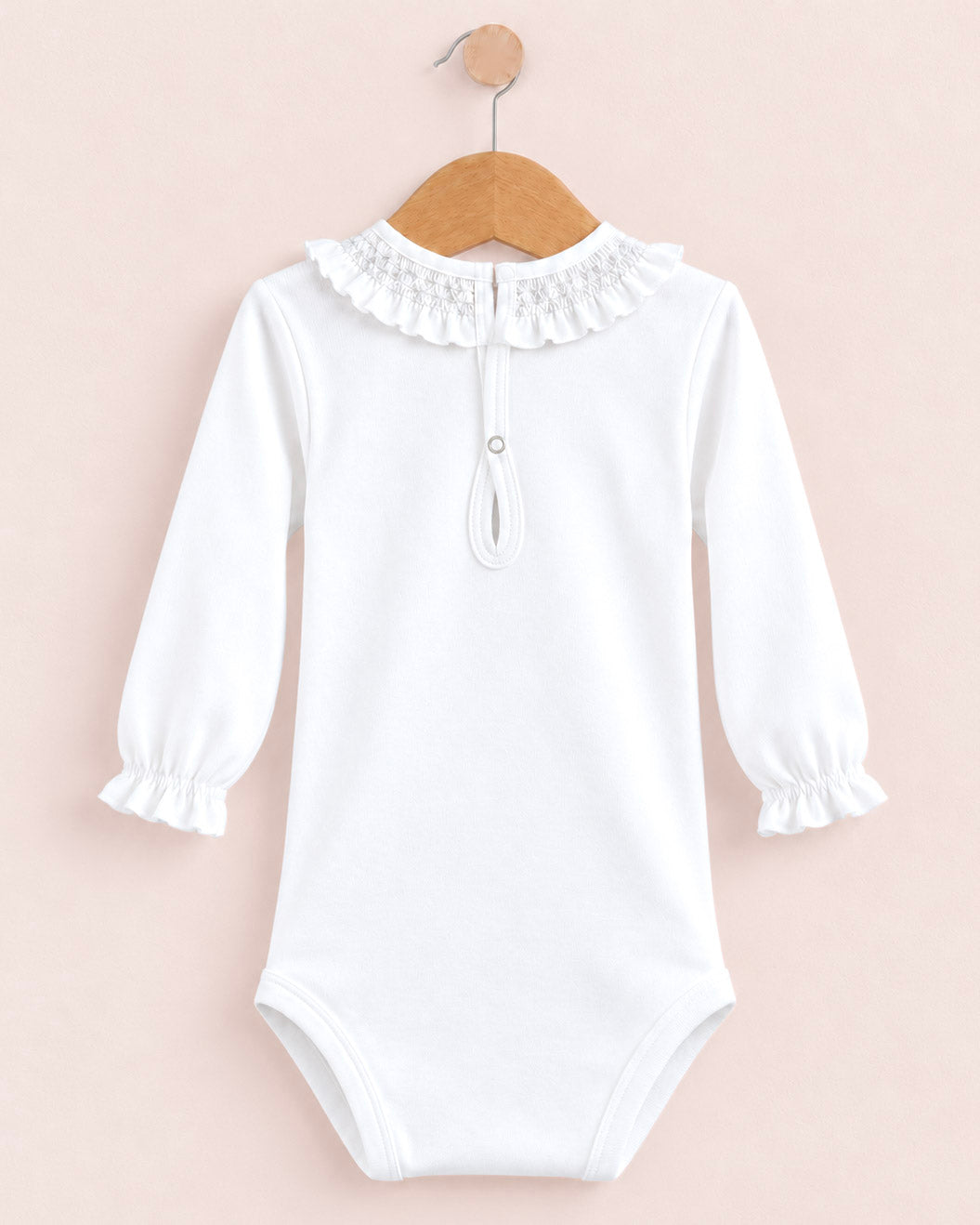 A white long-sleeve baby bodysuit with a ruffled lace collar and a small snap detail at the neck, hanging on a wooden hanger against a pale pink background.