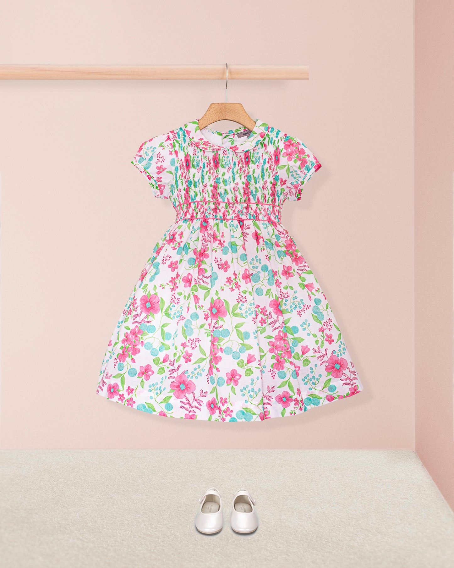 Alternate angled front view of the Amanda Palm Beach Floral Smocked Dress, displaying the smocked bodice, puff sleeves, and floral-printed full skirt on a hanger against a soft pink background styles with white Mary Jane shoes.
