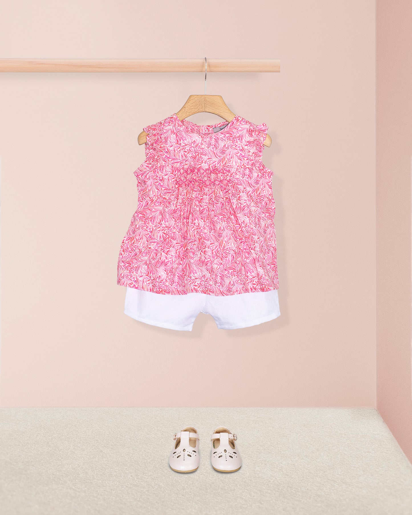 Amelie Liberty Wallace Garden Tunic styled with white shorts and white Mary Jane shoes, displayed against a pale pink wall and neutral carpet background.