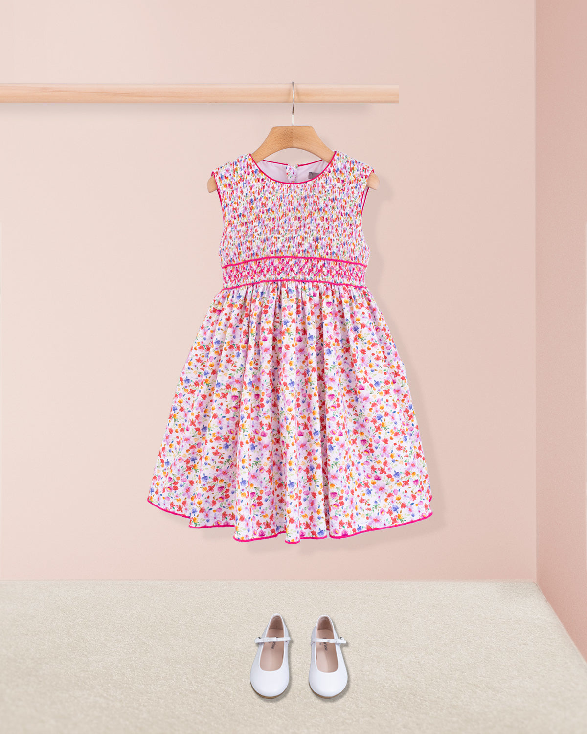 Amandine Veneto Hot Pink Garden Smock dress paired with white Mary Jane shoes, displayed simply on a neutral carpet against a pink background.