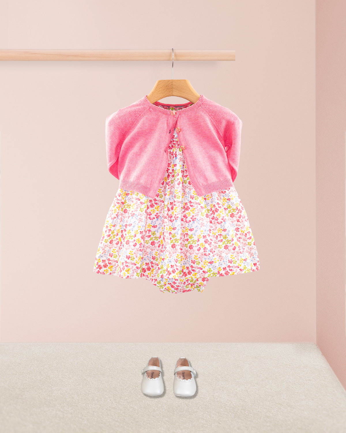 Baby Amelie floral dress styled with primrose pink cardigan and white Mary Jane shoes under a wooden rail