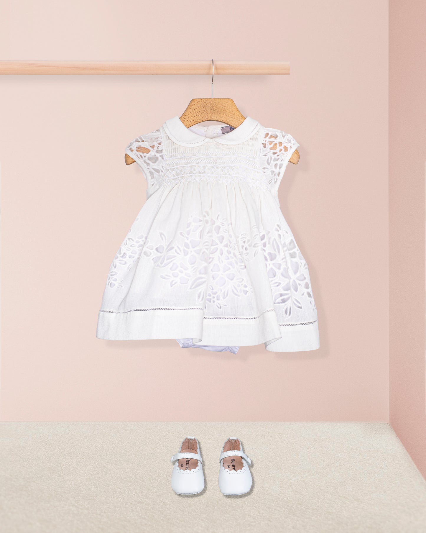 Styled shot of the Baby Emmy Doppia White Smock Dress Set with baby shoes, set against a soft peach background.