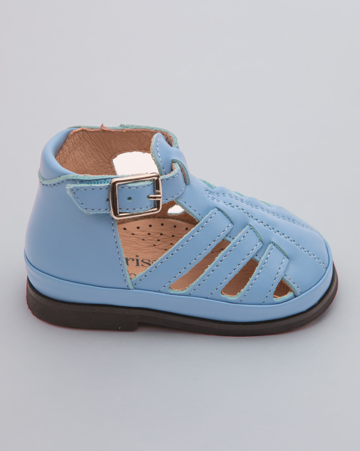 Side view of a light blue leather baby boy sandal with rubber sole, open side cutouts, and velcro ankle strap with decorative buckle, placed on a gray surface.