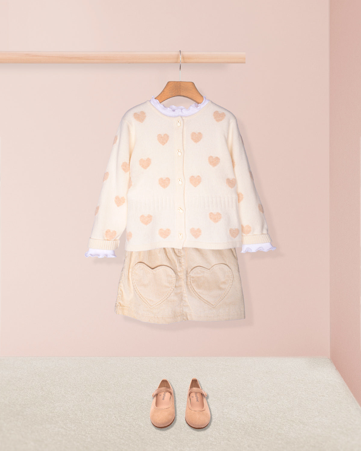 Children's outfit with heart-patterned top and skirt on a hanger against a pink wall.