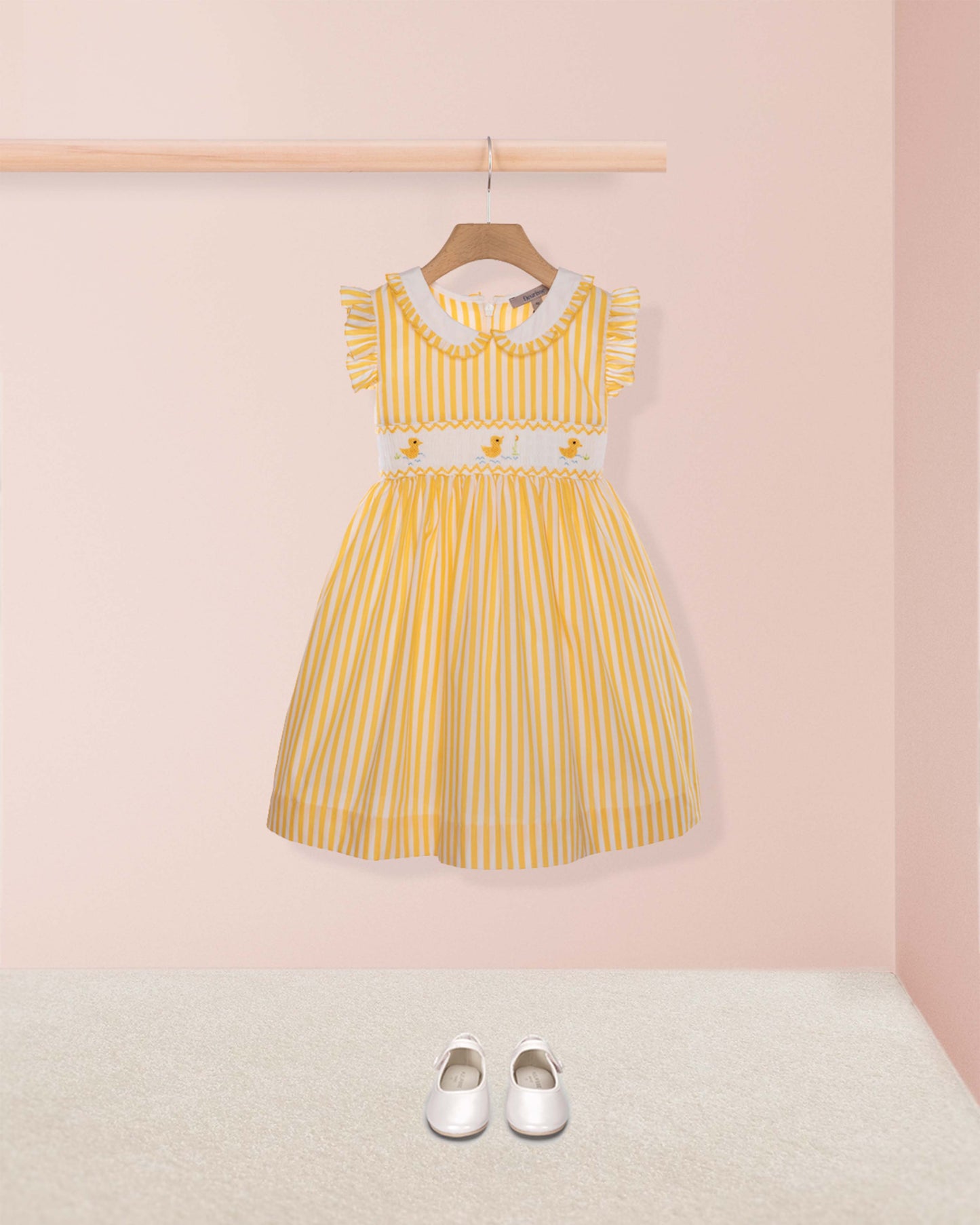Yellow striped dress on a hanger against a pink wall with white shoes on the floor.