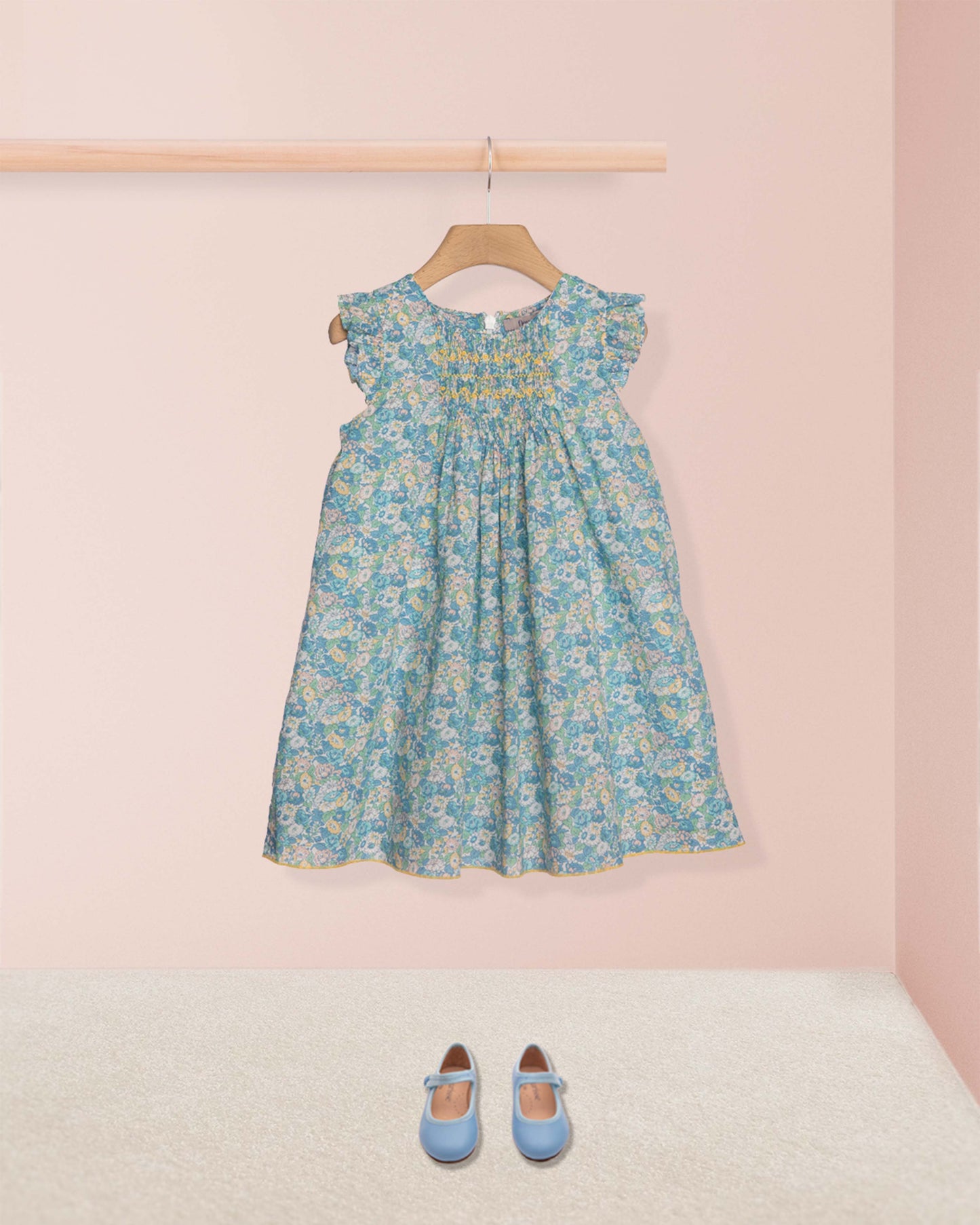 Floral dress on a hanger with matching shoes on the floor against a pink wall.