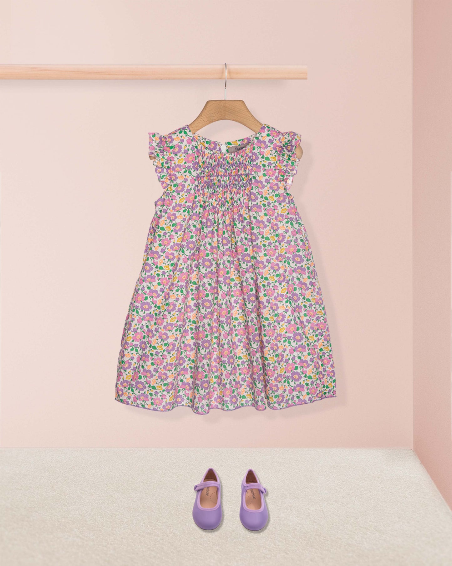 Floral dress on a hanger with matching shoes on the floor against a pink wall.
