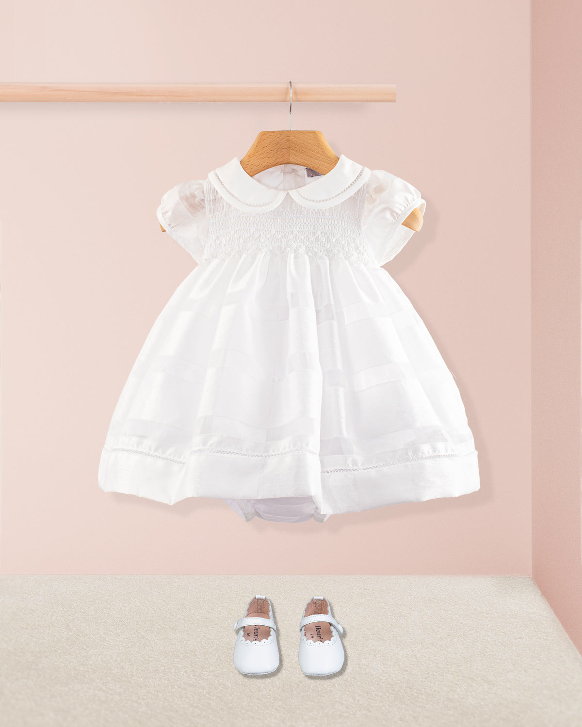 Baby Emmy White Chiffon Dress styled with soft white crib shoes placed on the floor below. The dress hangs on a wooden rod against a blush pink background with neutral carpet underneath.