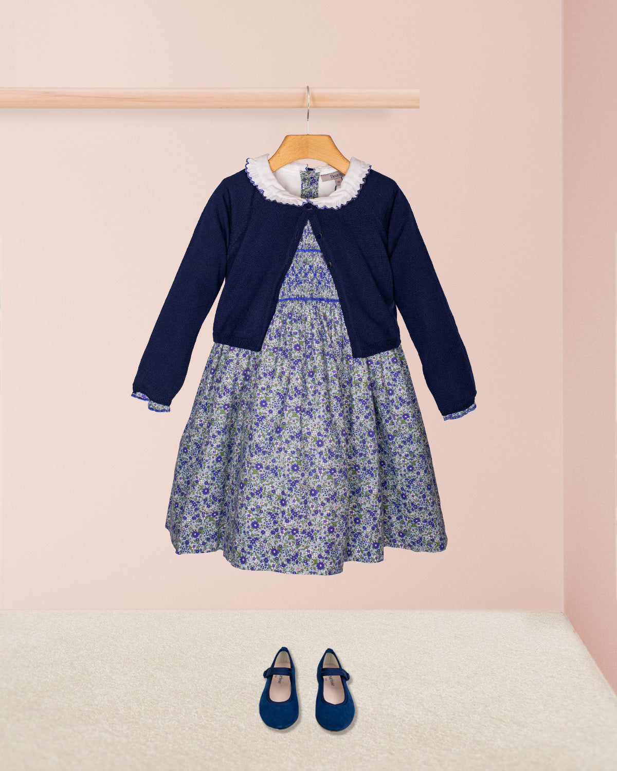The Fiorella Belina dress styled with the Serena Cardigan in a navy knit and paired with navy blue suede Mary Jane shoes, displayed against a pastel pink backdrop.