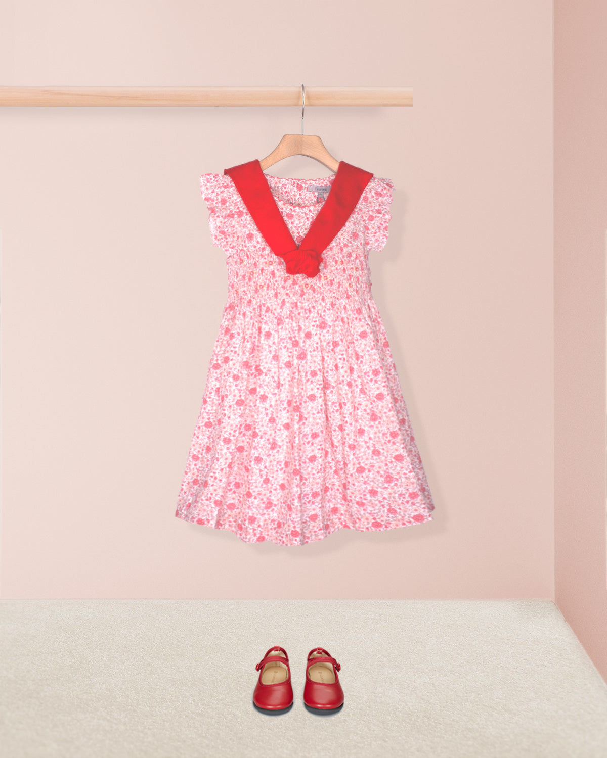 Pink floral dress with red trim on a hanger against a pink wall, with matching shoes on the floor.