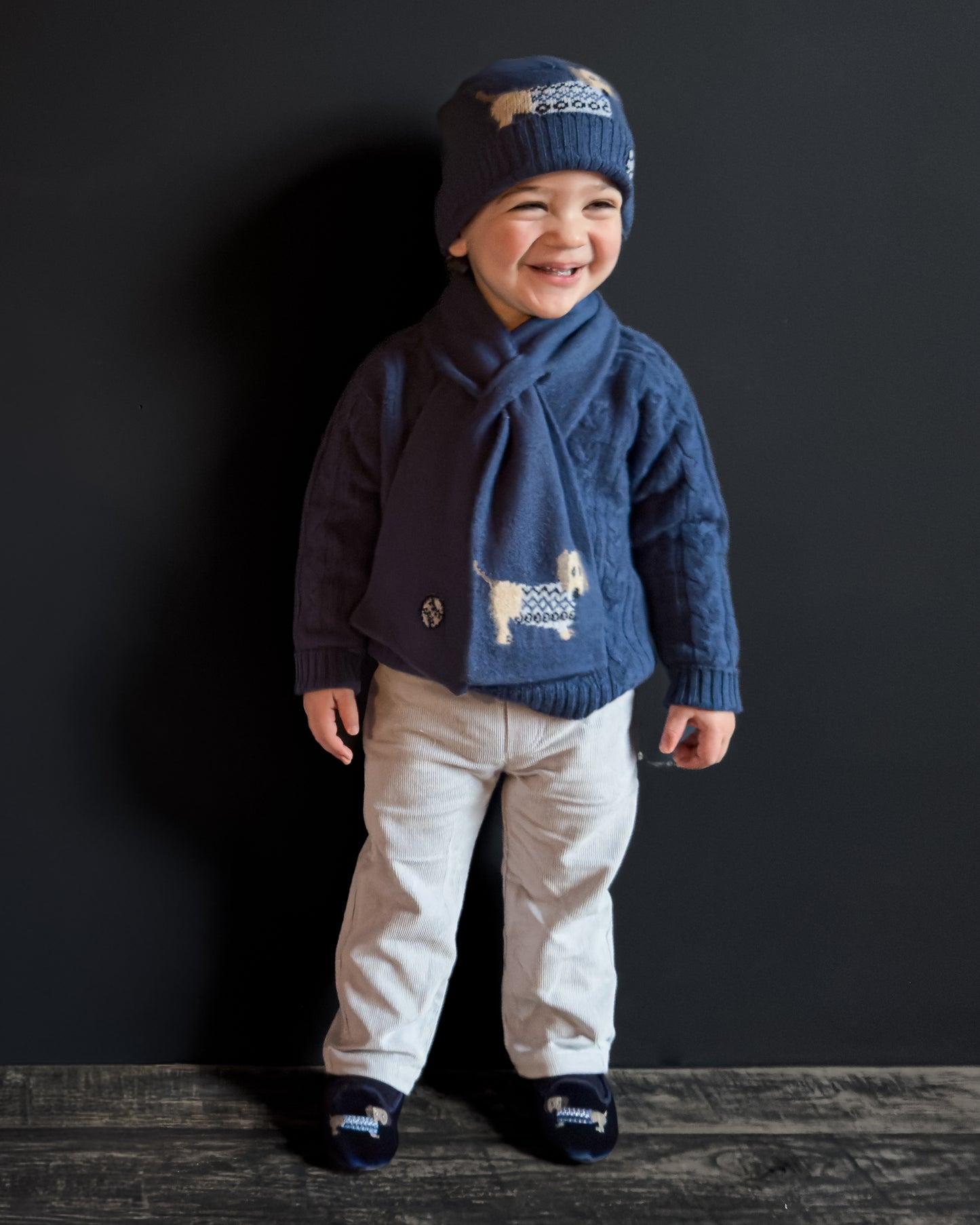 Child wearing a blue sweater with a dog design, beige pants, and a matching hat on a dark background