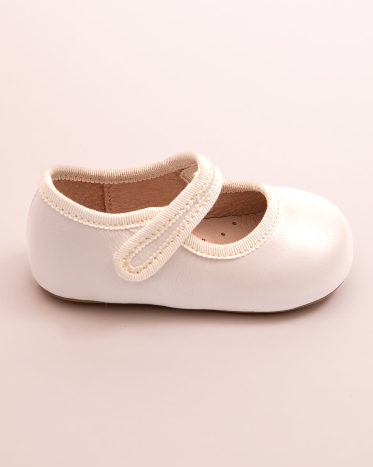 Side view of a cream leather baby Mary Jane shoe with a Velcro strap and soft sole, shown on a pink surface.