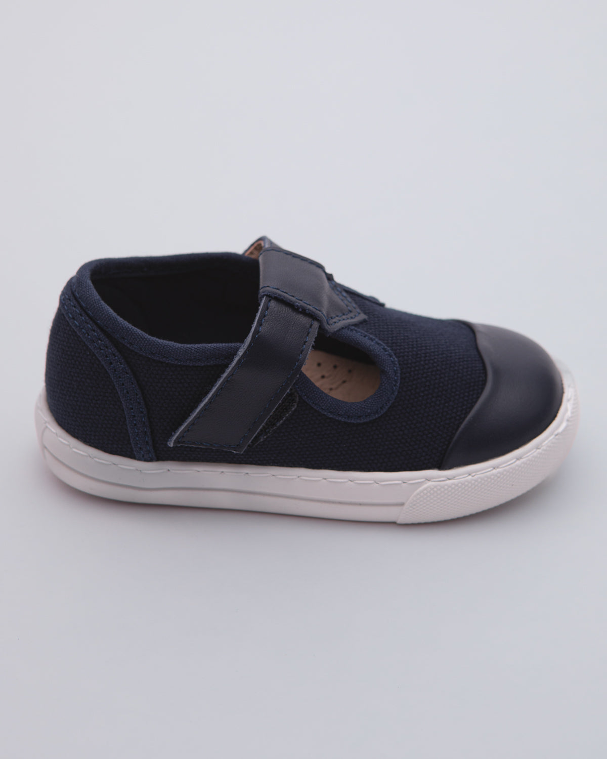 Side view of a navy blue toddler sneaker showing the white rubber sole, canvas body, leather toe cap, and velcro strap closure on a gray surface.