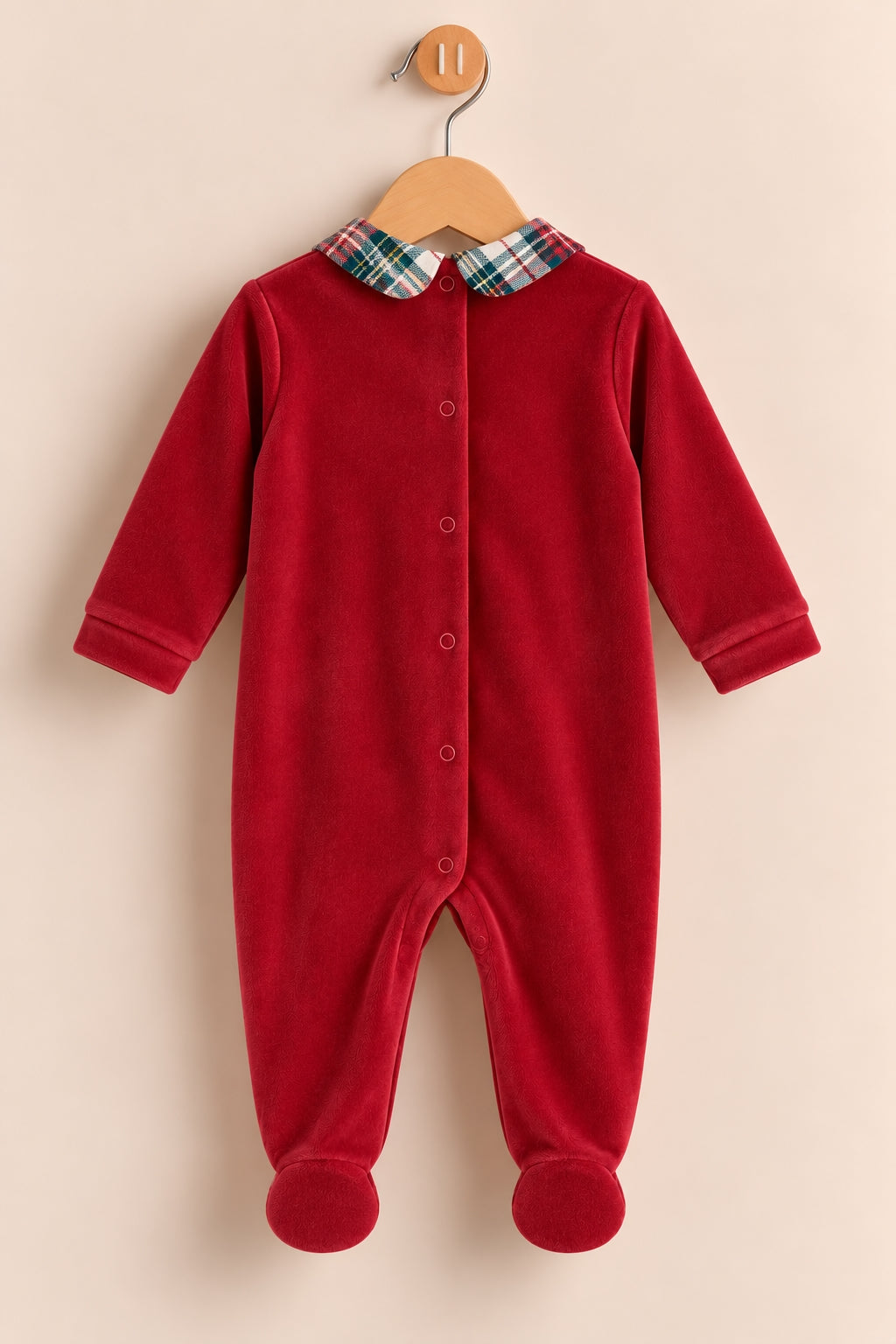 A baby's red footed sleeper with a plaid collar, hanging on a wooden hanger against a light beige background.