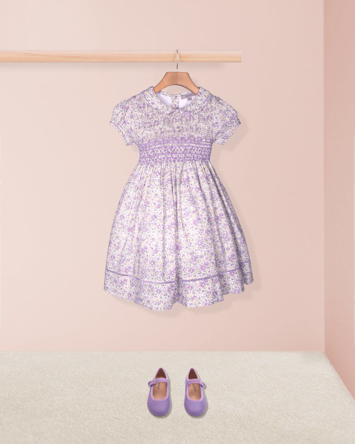 Lavender floral dress on a hanger with matching shoes on the floor against a pink wall.