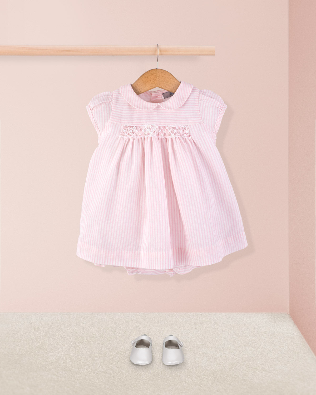 Front view of Baby Eva 2-piece dress paired with silver crib shoes, displayed hanging on a wooden rod above a carpeted floor for a full styled look.