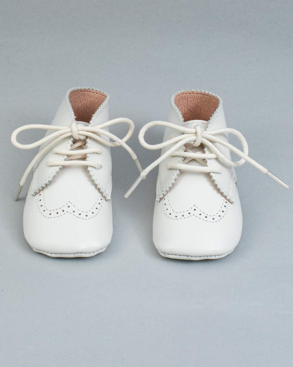 Front view of a pair of cream leather crib shoes with tied laces and scalloped brogue details.