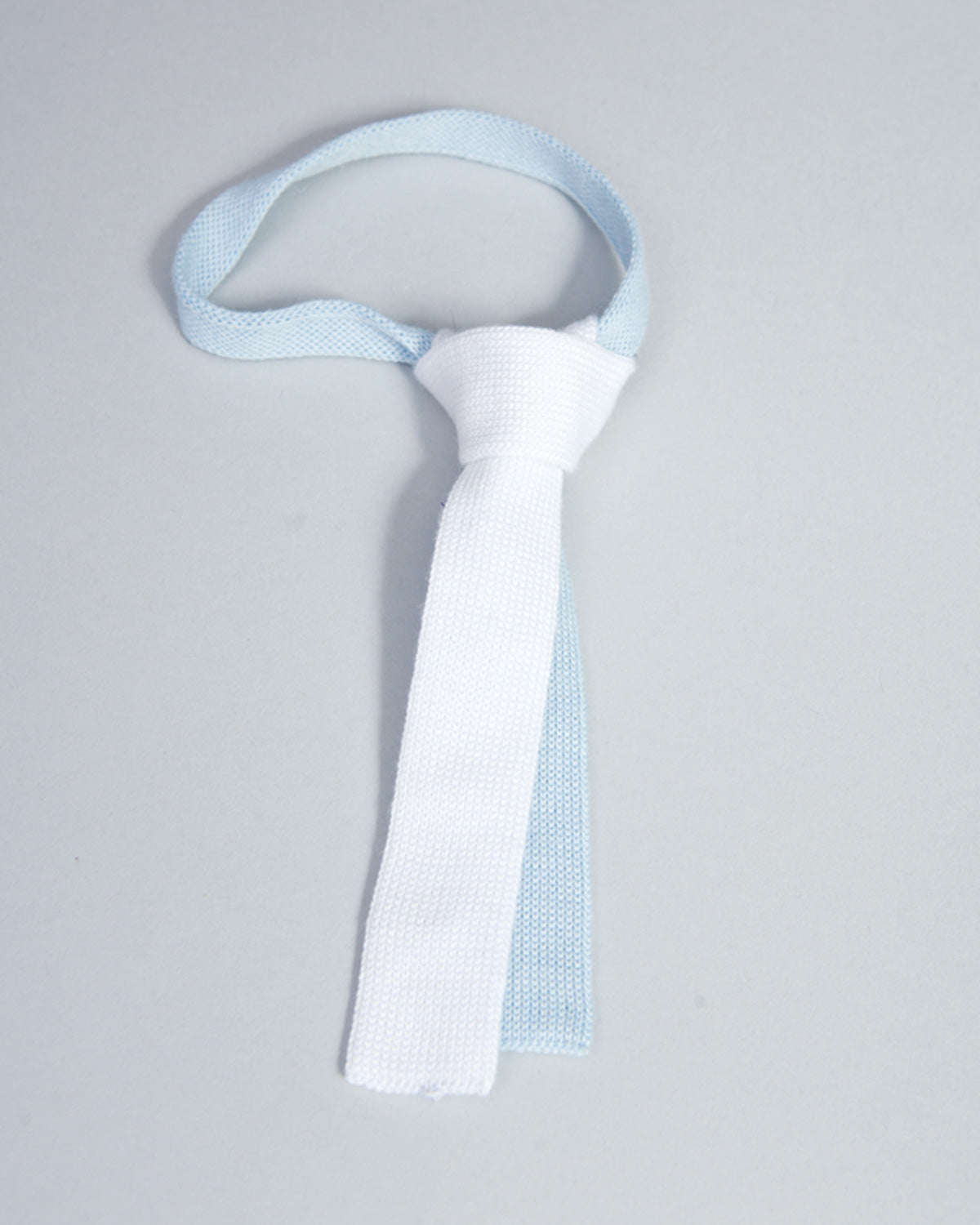 White and light blue Italian cotton knit baby tie displayed against a soft gray background.