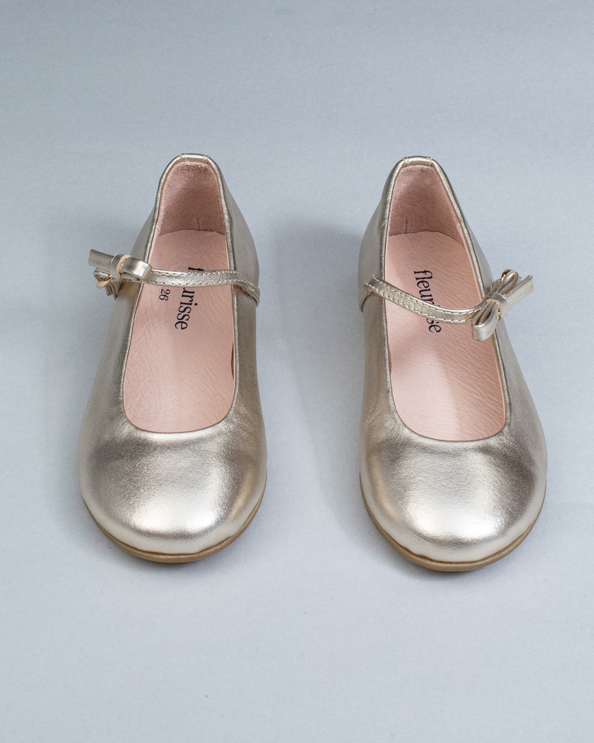 Frontal view of gold leather Mary-Jane shoes for children, showcasing symmetrical bows and rounded toes on a flat background.