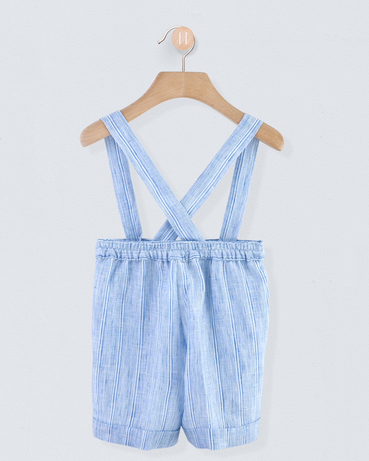 Back view of blue striped linen suspender shorts with crossed straps and elastic waistband, shown on a wooden hanger