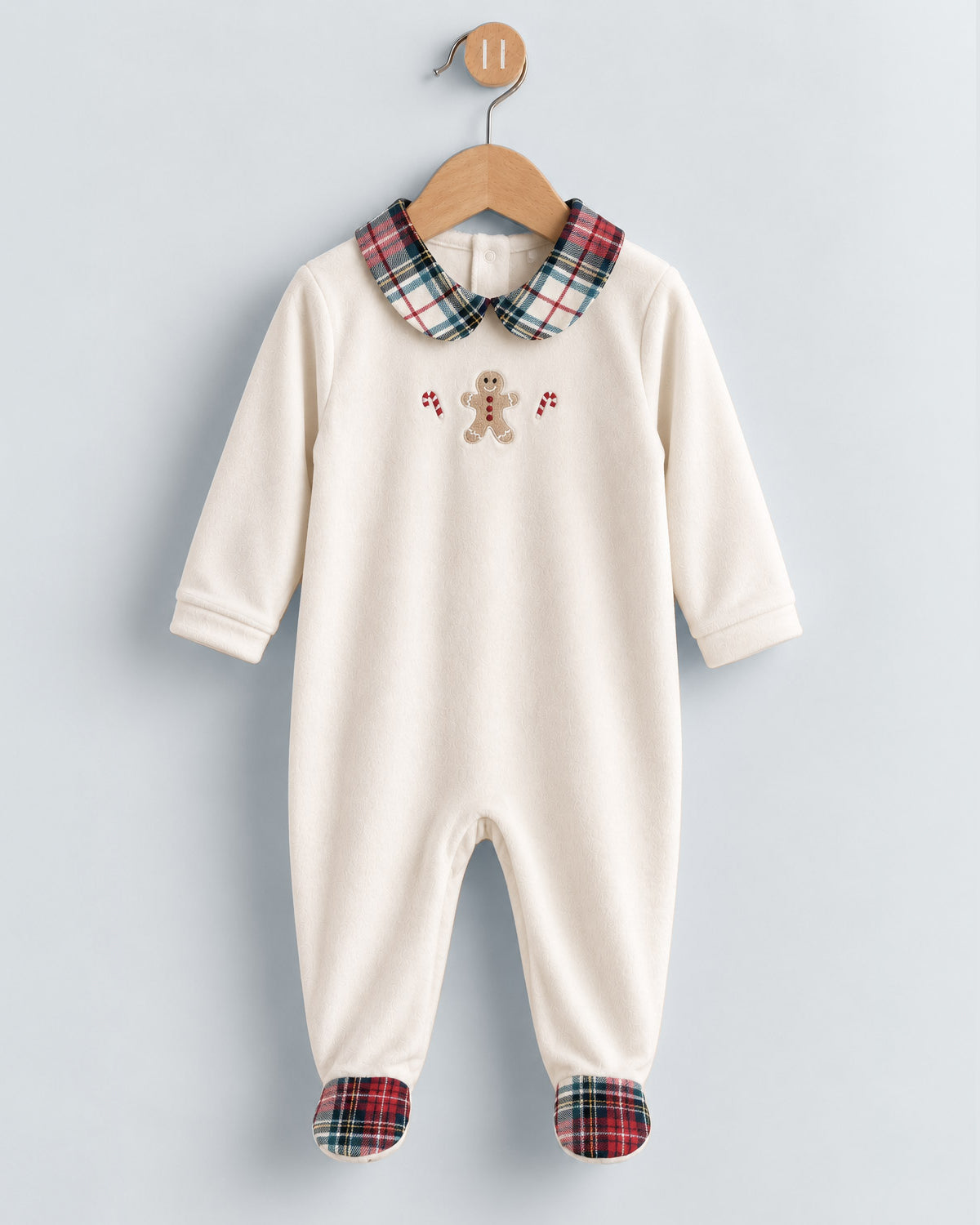 A baby footed one-piece jumpsuit in off-white, featuring a red, green, and blue plaid Peter Pan collar and matching plaid toe caps, with a small gingerbread man embroidery on the chest, hanging on a wooden hanger against a pale blue background.