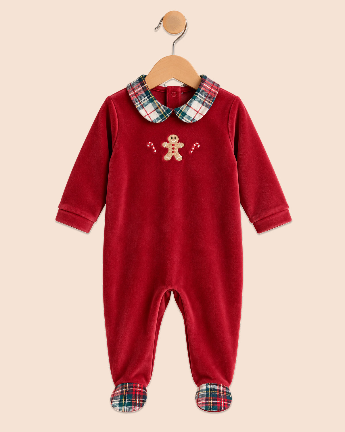 A baby footed one-piece jumpsuit in deep red velvet, featuring a green and red plaid collar and matching plaid cuffs, with a small gingerbread man embroidery on the chest. The garment is displayed on a wooden hanger against a solid beige background.