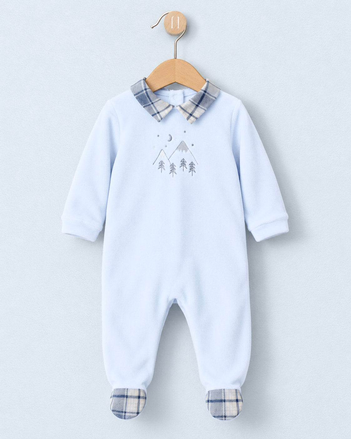 A baby footed one-piece jumpsuit in light blue, hanging on a wooden hanger against a light gray background. The jumpsuit features a plaid collar and matching plaid foot cuffs, with a small gray mountain and tree graphic on the chest.