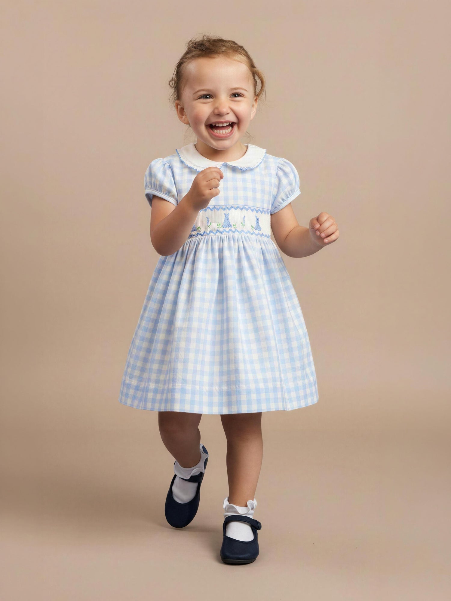 Little girl wearing a light blue checkered dress with a strip of bunnies across the chest, and black shoes with short socks, against a beige studio backdrop