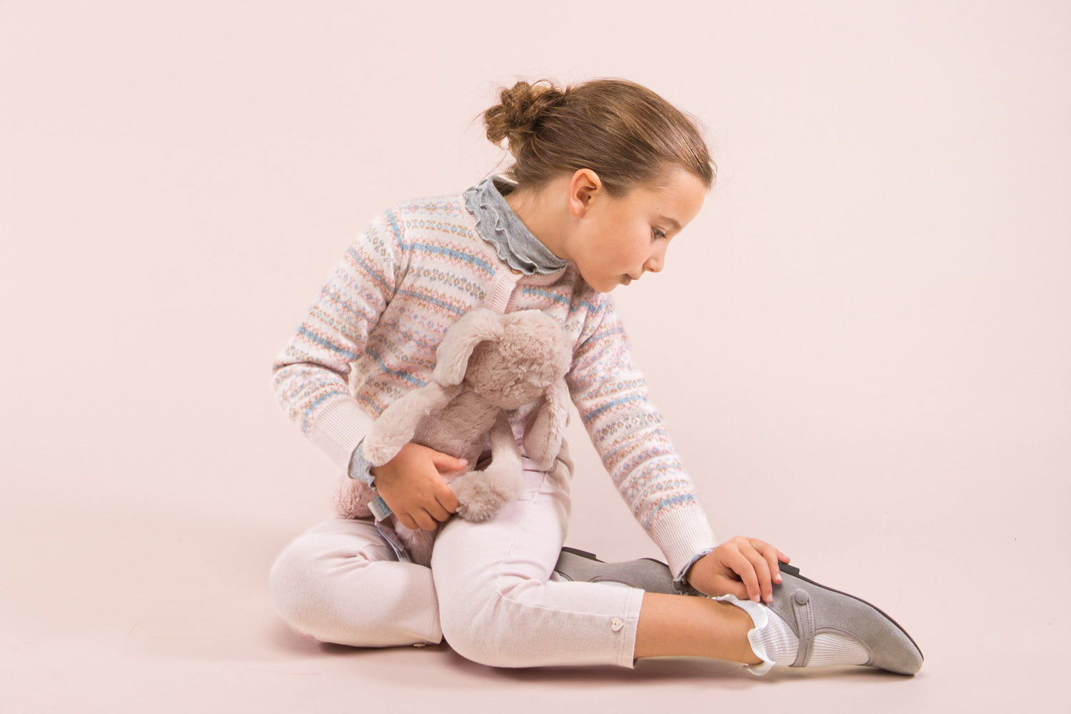girl sitting on floor wearing pink stripe cashmere cardigan, pink leggings and grey suede shoes holding bunny plush toy