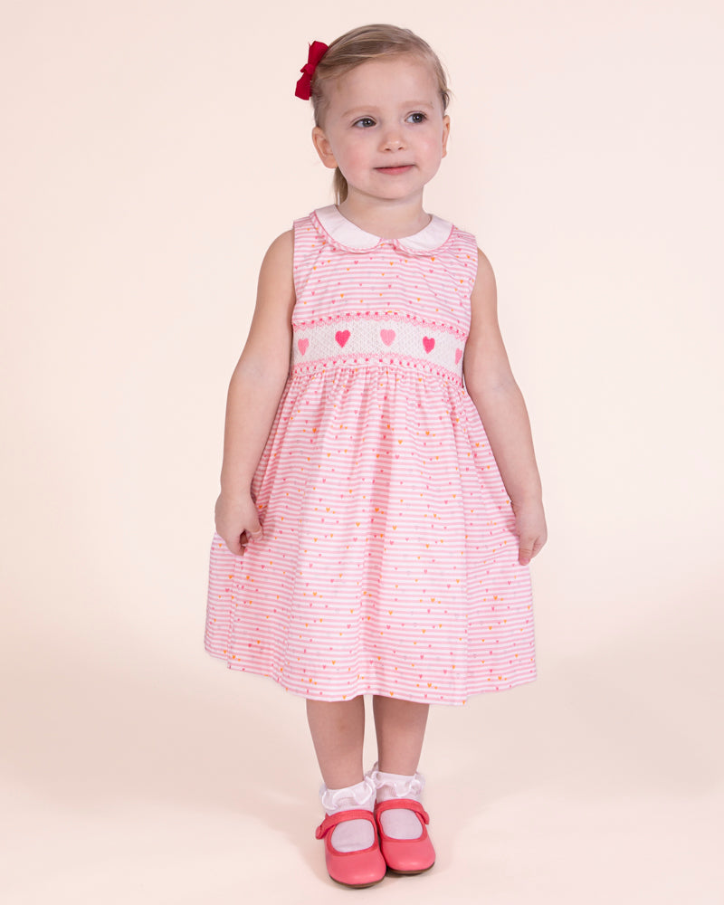 Girl model standing, wearing Emma Delicate Blush Rose Smock Dress showing the front of the dress with a Pink bag on the floor to match the outfit 