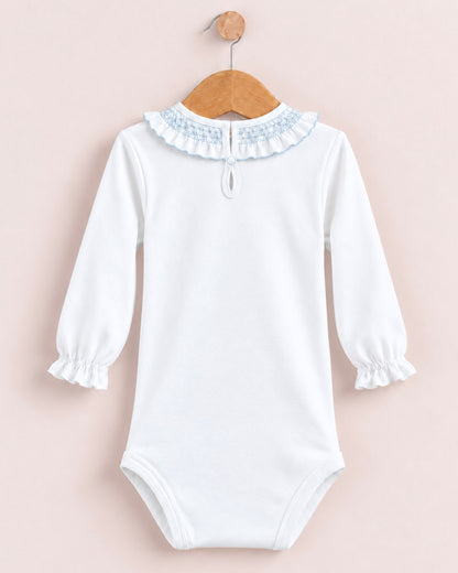 A white long-sleeve baby bodysuit with a light blue smocked ruffled collar, hanging on a wooden hanger against a pale pink background.