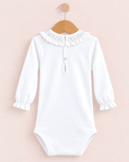 A white long-sleeve baby bodysuit with a ruffled lace collar and a small snap detail at the neck, hanging on a wooden hanger against a pale pink background.