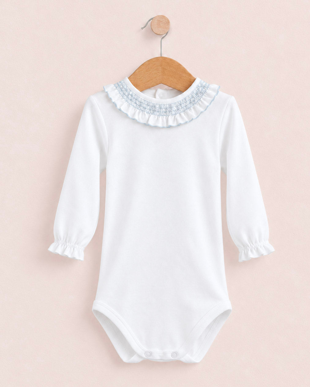 A white long-sleeve baby bodysuit with a ruffled collar and smocked cuffs, hanging on a wooden hanger against a soft pink background.