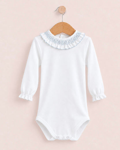 A white long-sleeve baby bodysuit with a ruffled collar and smocked cuffs, hanging on a wooden hanger against a soft pink background.