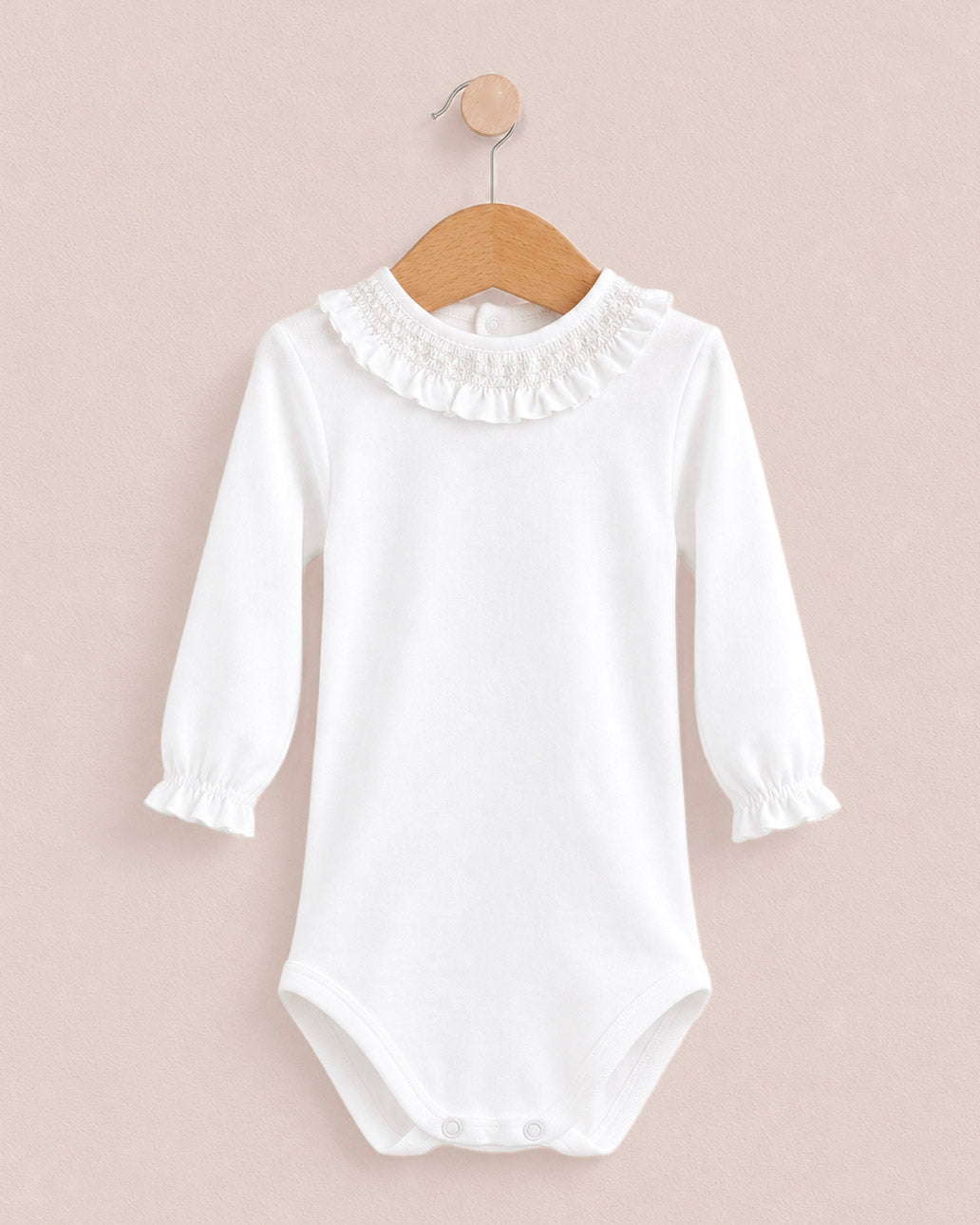 A white long-sleeve baby bodysuit with a ruffled collar and elastic cuffs, hanging on a wooden hanger against a solid pale pink background.