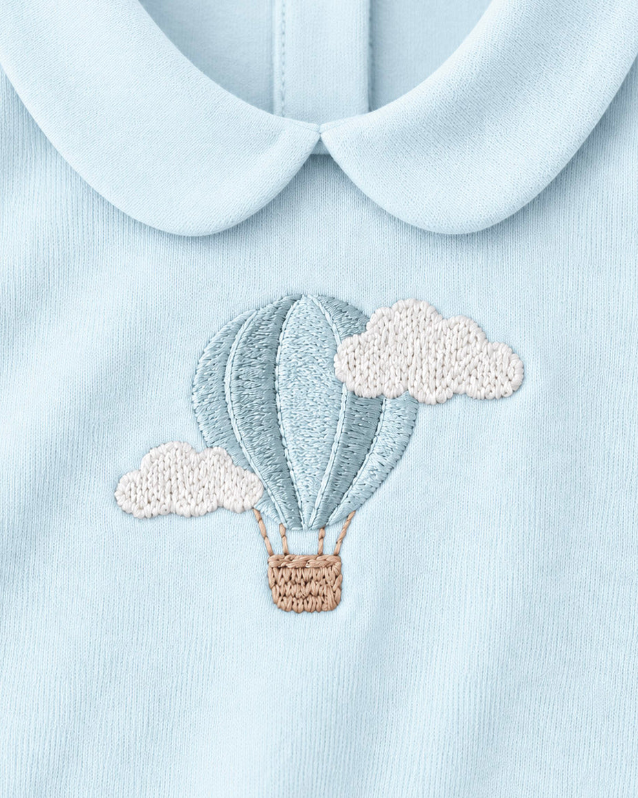 Close-up of a baby garment with a light blue Peter Pan collar and an embroidered hot air balloon motif in matching blue, cream, and tan on the chest.