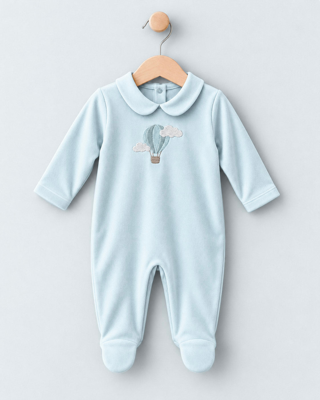 A baby long-sleeve footed jumpsuit in light blue, featuring a cute hot air balloon and cloud graphic on the chest, hanging on a wooden hanger against a light gray background.