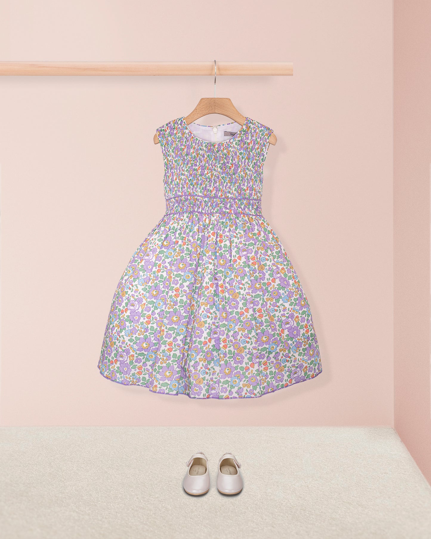 Front view of the Amandine Liberty Betsy Lavender Smock dress displayed with white Mary Jane shoes on a carpeted floor, against a light pink wall.