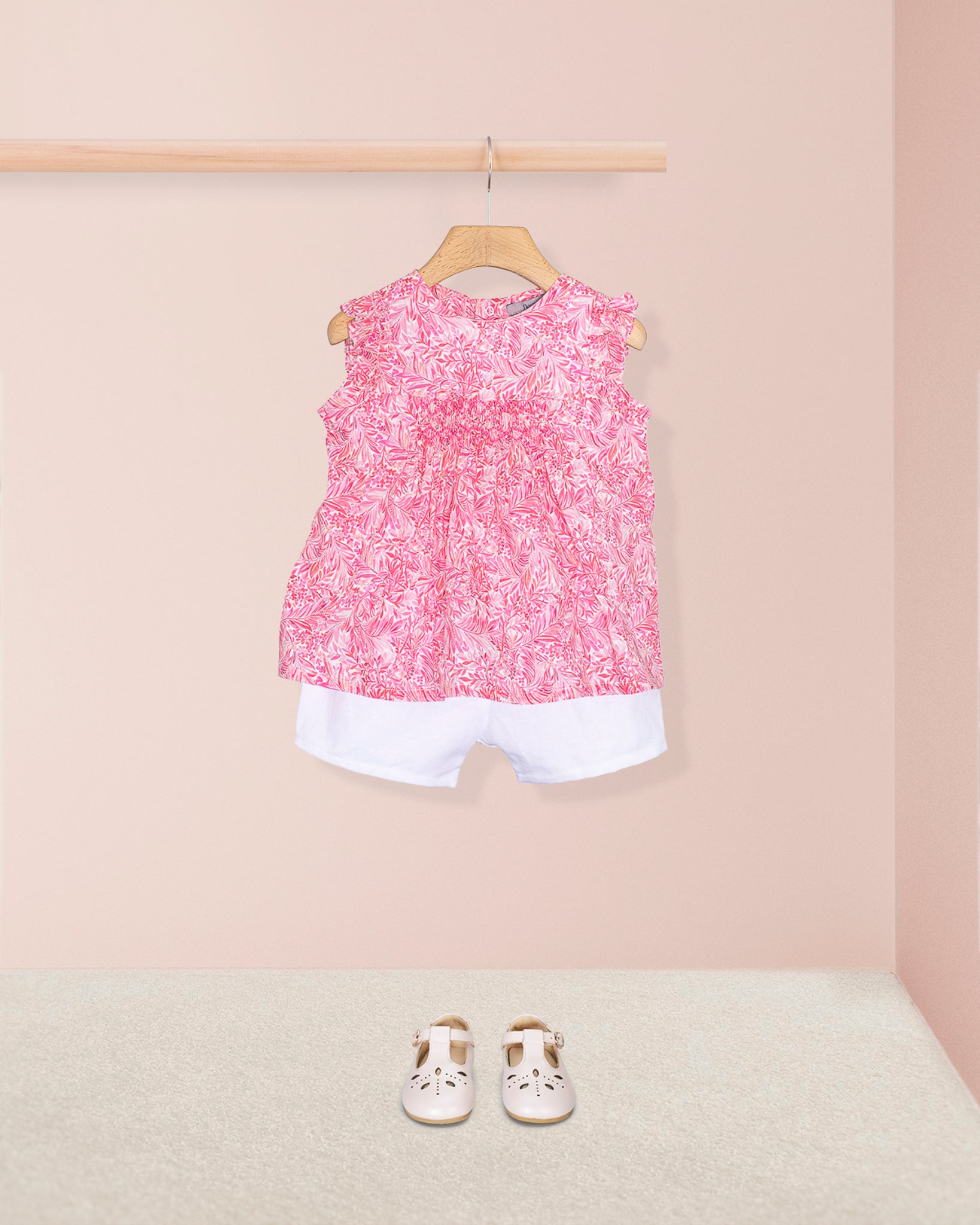 Amelie Liberty Wallace Garden Tunic styled with white shorts and white Mary Jane shoes, displayed against a pale pink wall and neutral carpet background.