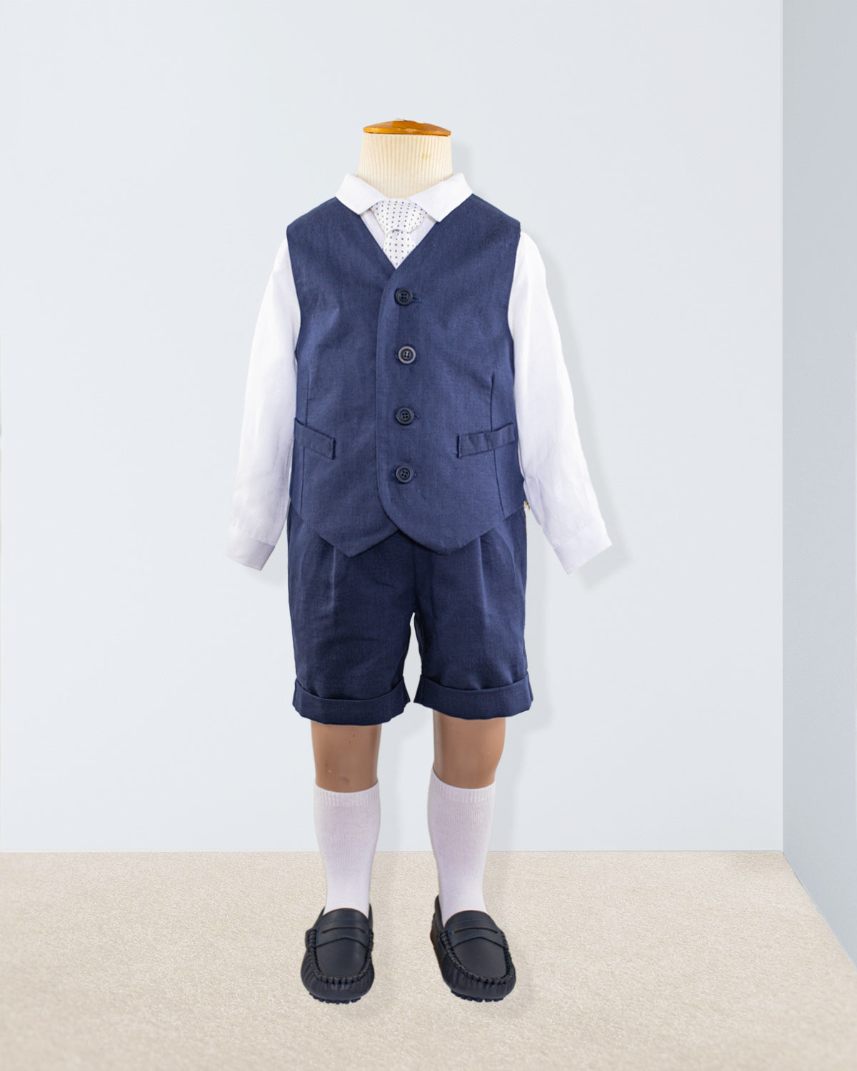 Navy linen suspender shorts and vest styled over a white long-sleeve shirt, shown on a child-sized mannequin with knee-high socks and navy shoes