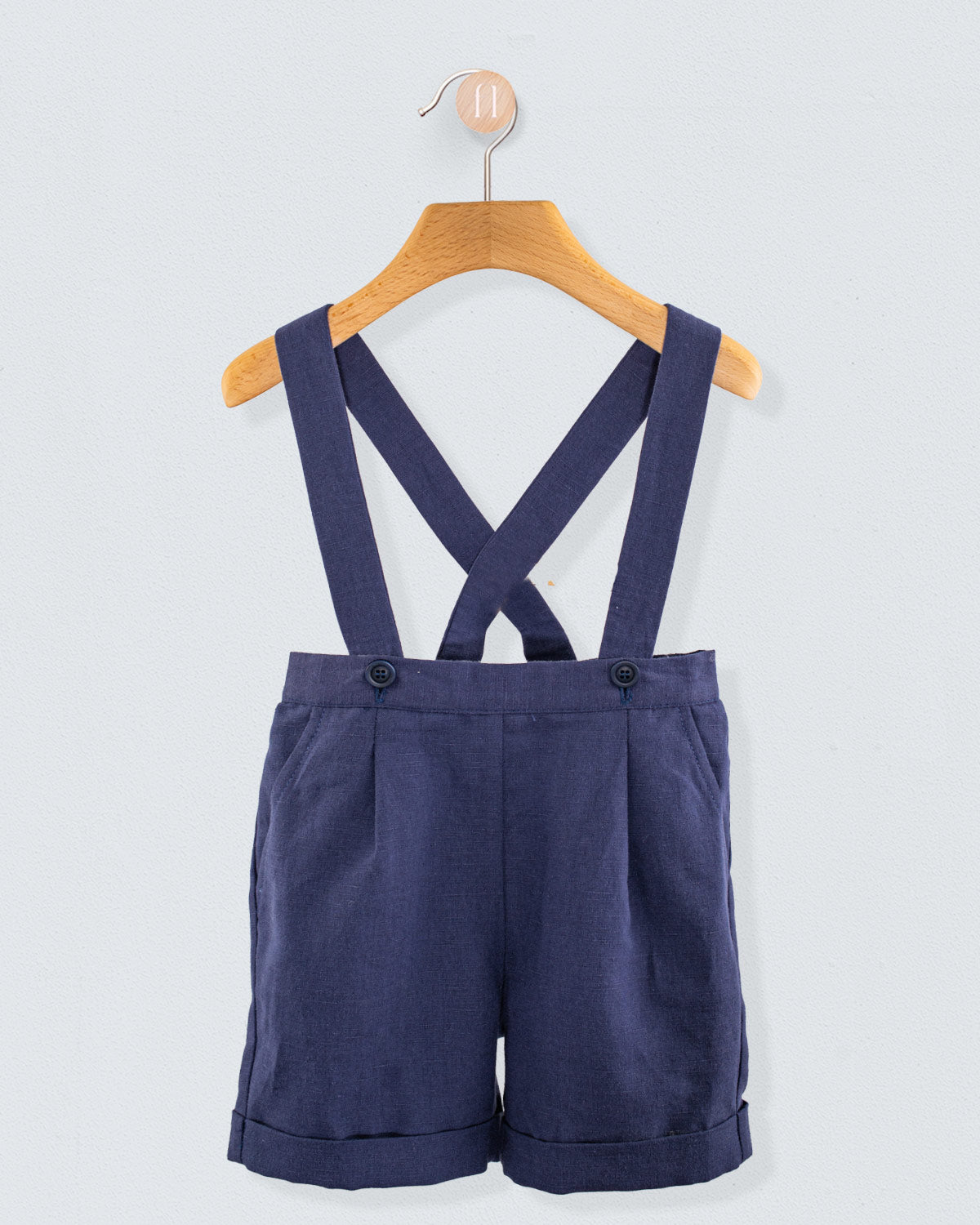 Front view of navy linen suspender shorts with crossed straps and waistband button detail, displayed on a hanger