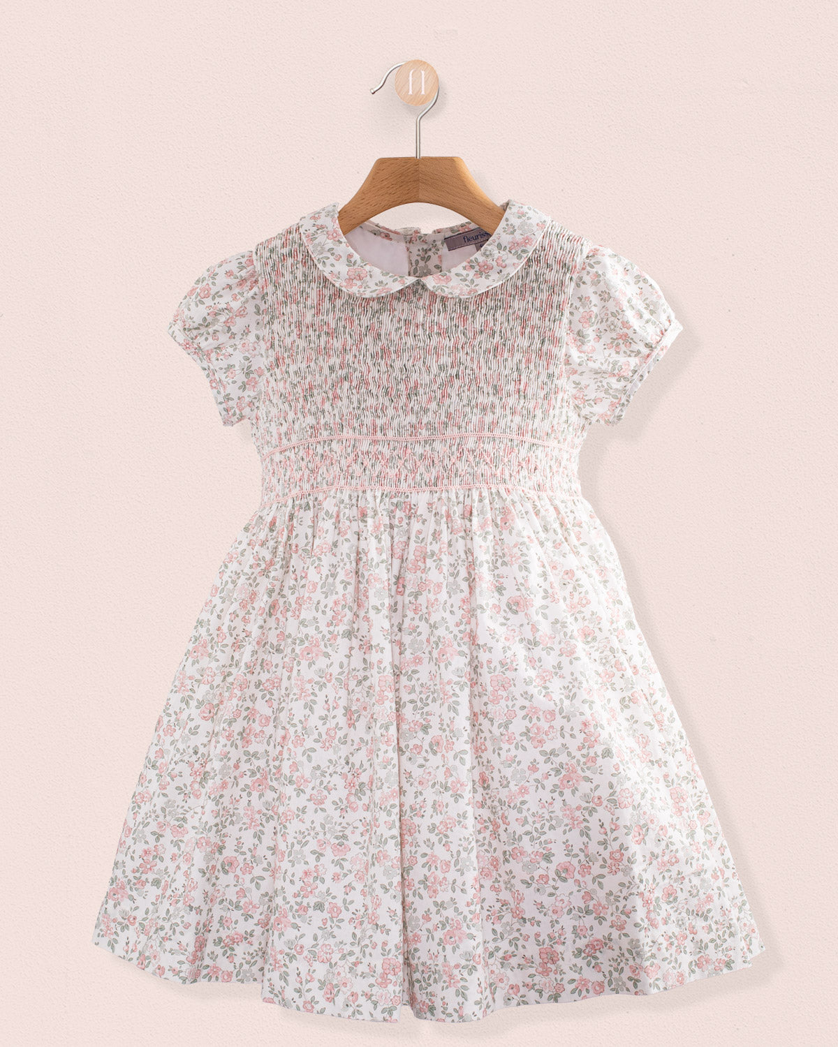 Front view of Amanda Blush Garden Smocked Dress hanging on a wooden hanger against a pink wall