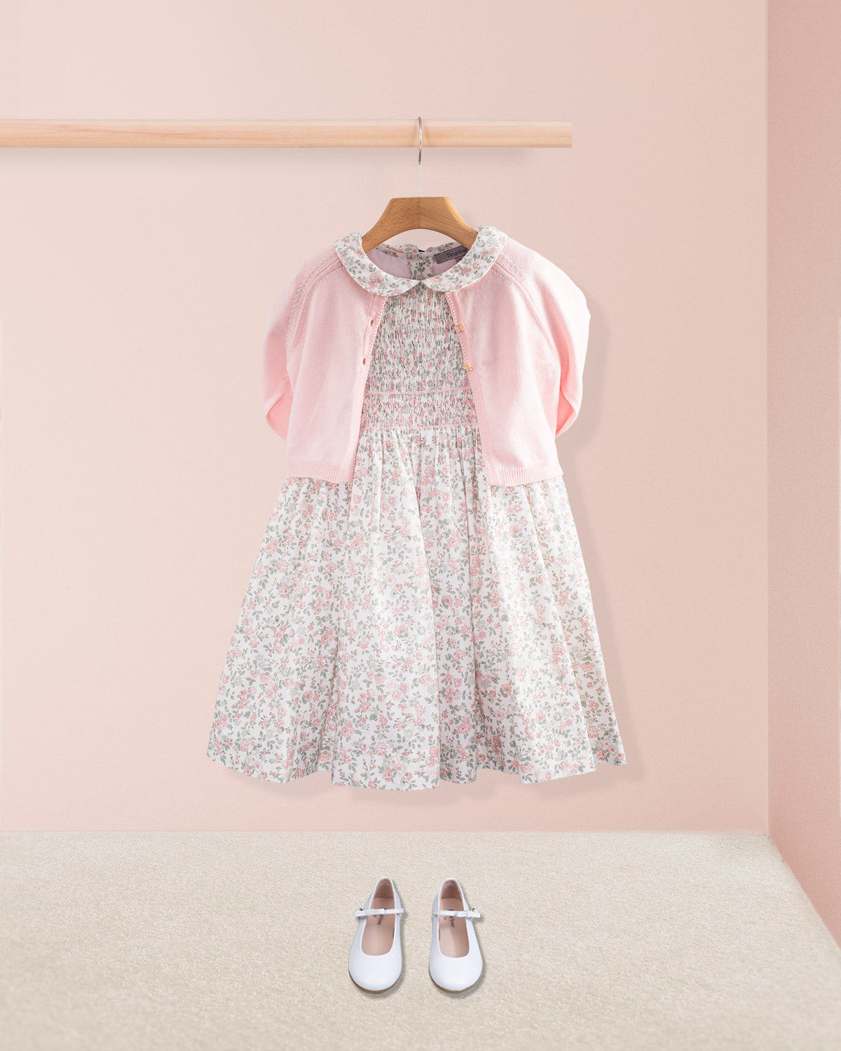 Amanda Blush Garden Smocked Dress styled with a pink cardigan and white shoes on a pink wall background