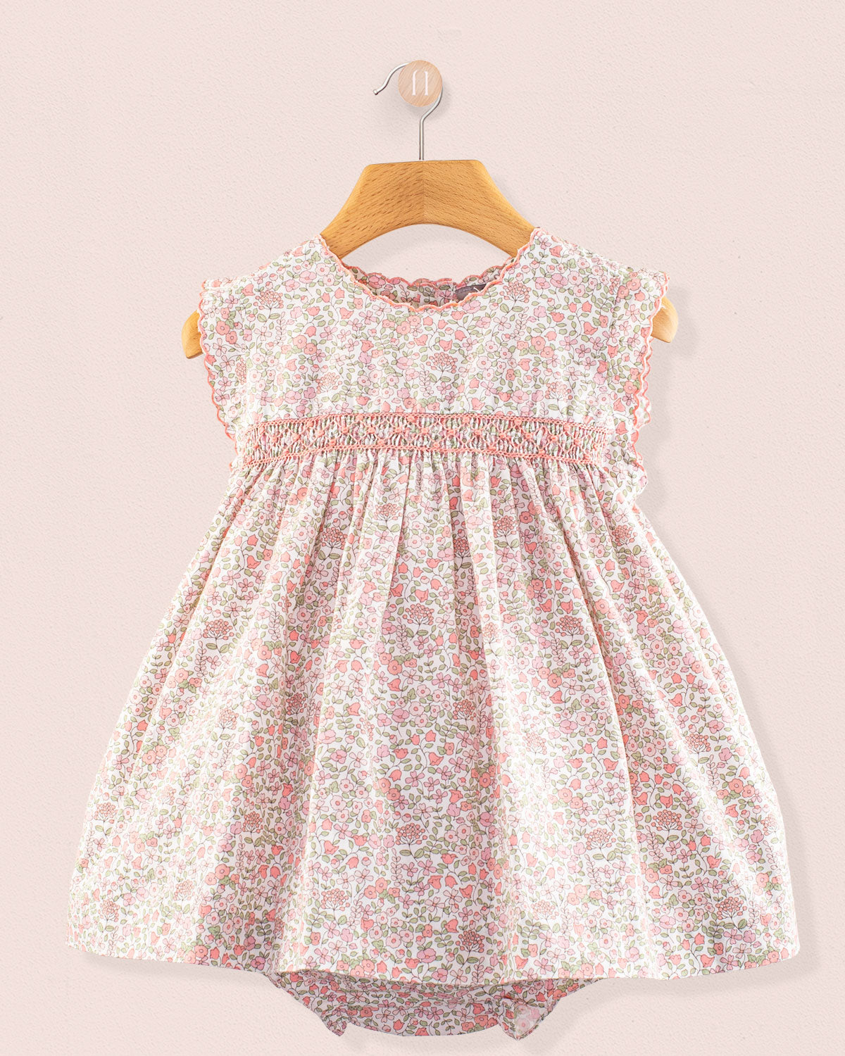 Baby Amelie Garden Pink Set front view featuring a pink floral smocked dress with cap sleeves and gathered skirt on a wooden hanger against a pale pink wall