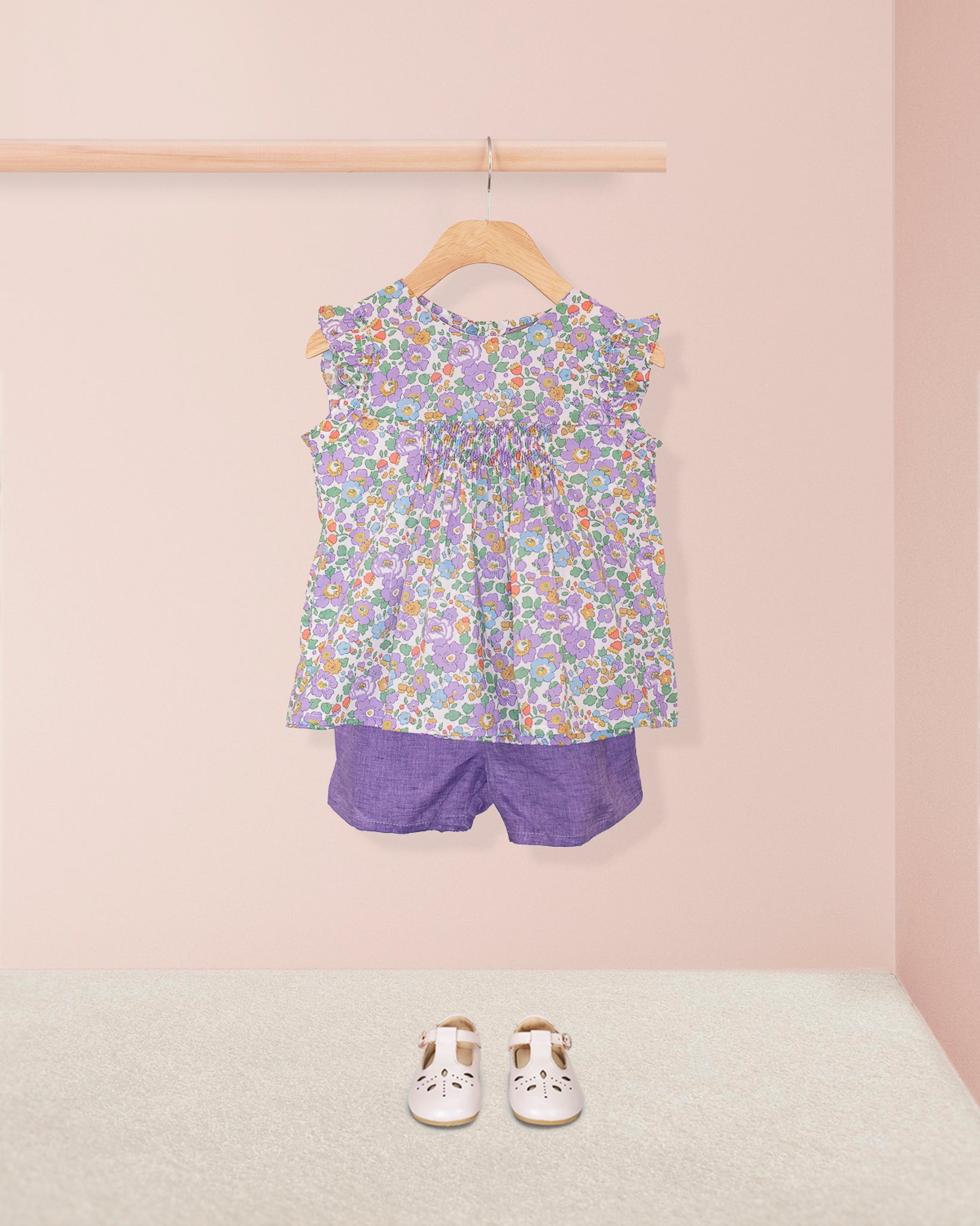 Amelie Liberty Betsy Lavender Tunic styled with purple shorts and white perforated shoes, displayed on a neutral carpet with a soft pink wall background.
