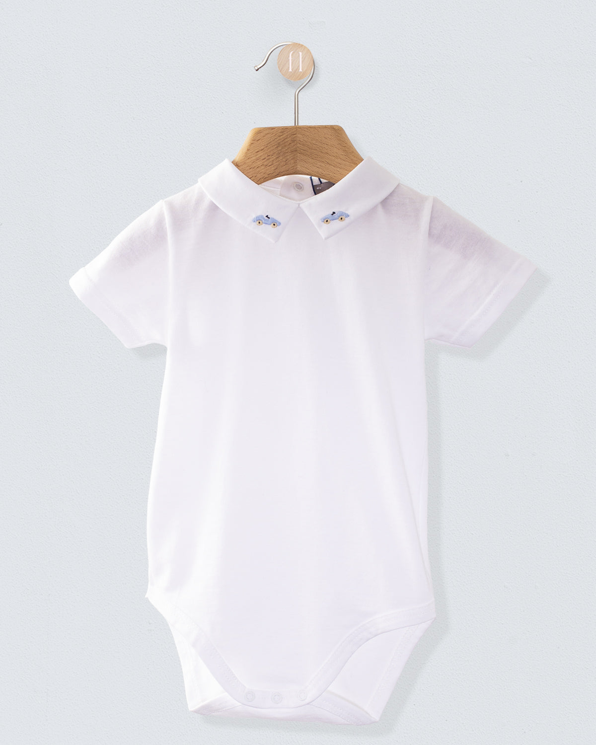 White baby onesie made from fine Italian cotton, displayed on a wooden hanger, with a collar embroidered with light blue car motifs on both sides.