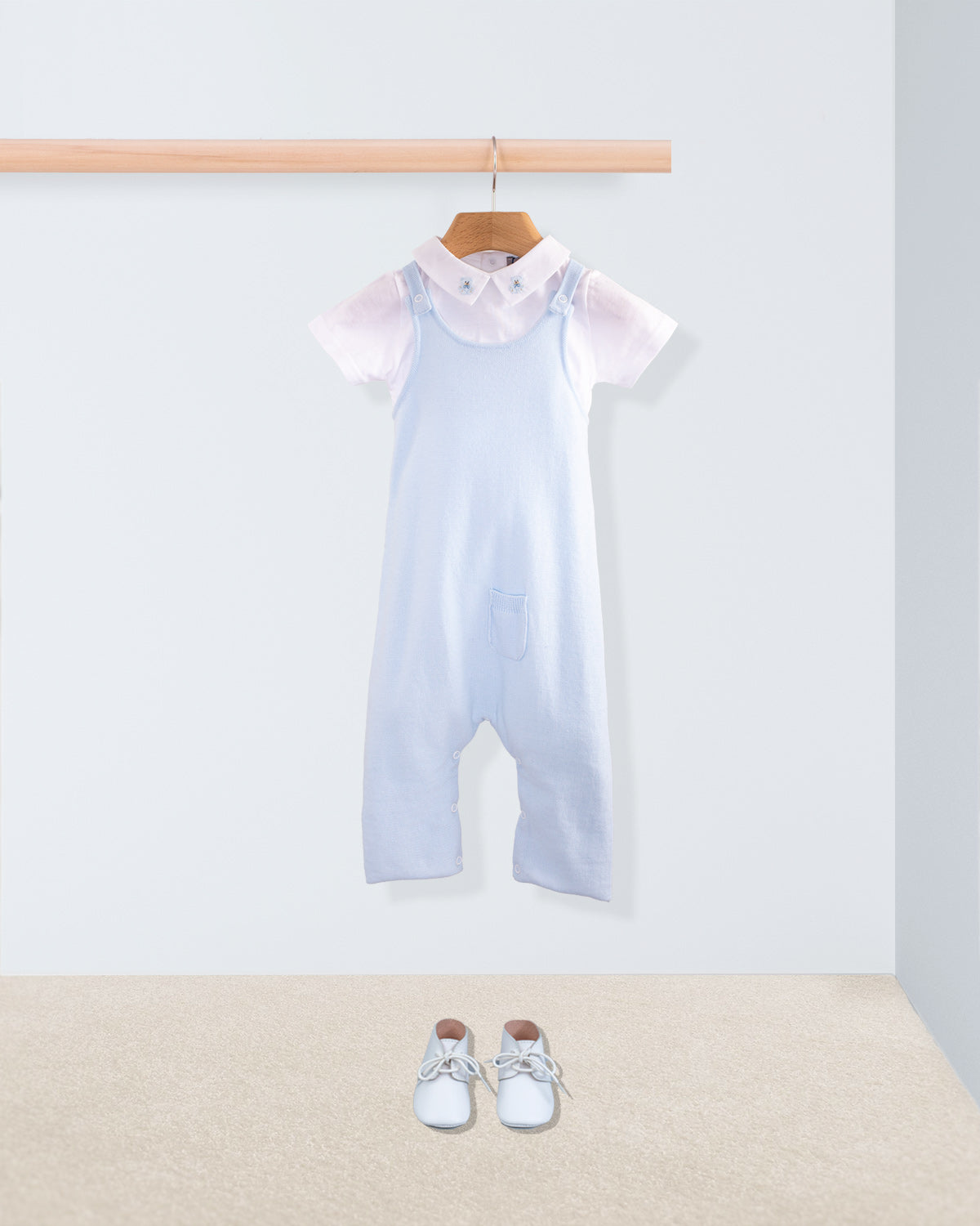 Aspen Powder Blue Cotton Knit Overall styled over a short-sleeve white embroidered-collar onesie, shown with a pair of white baby shoes on a beige carpet for a complete look.