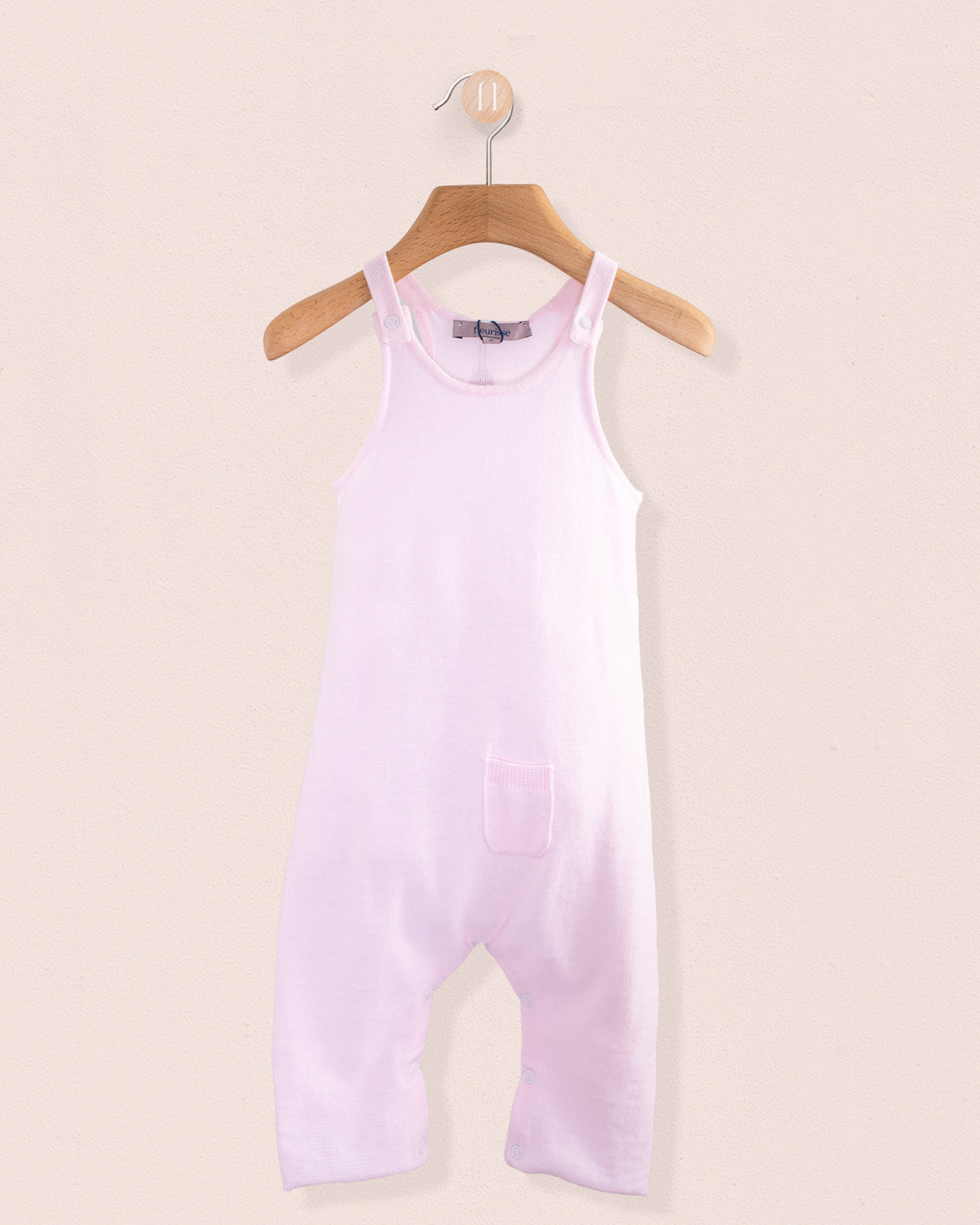 Light pink cotton knit baby overall hanging on a wooden hanger against a pale pink background