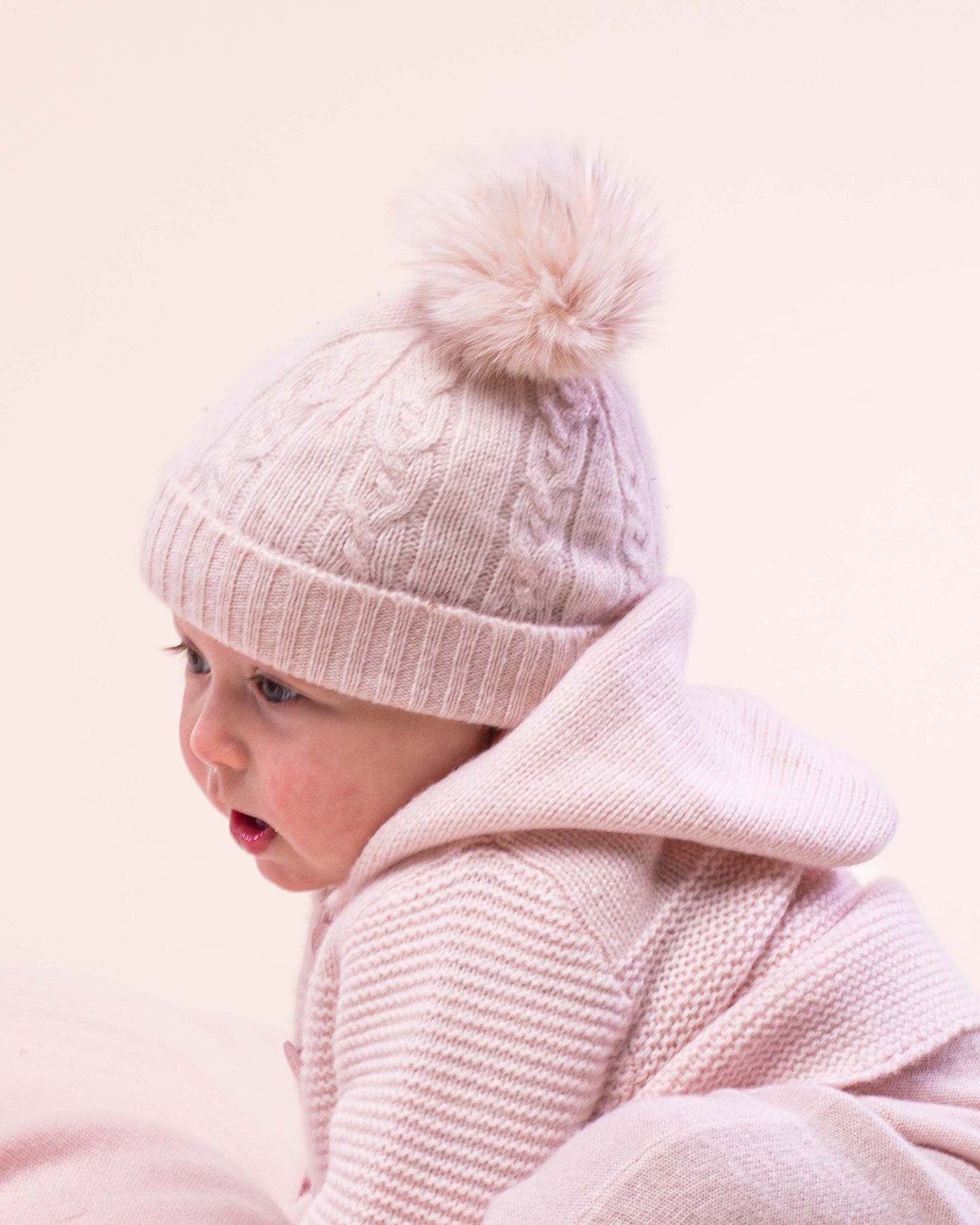 Baby girl wearing the Cashmere Pink Fur Pom-Pom Hat facing sideways to show the details of the hat 