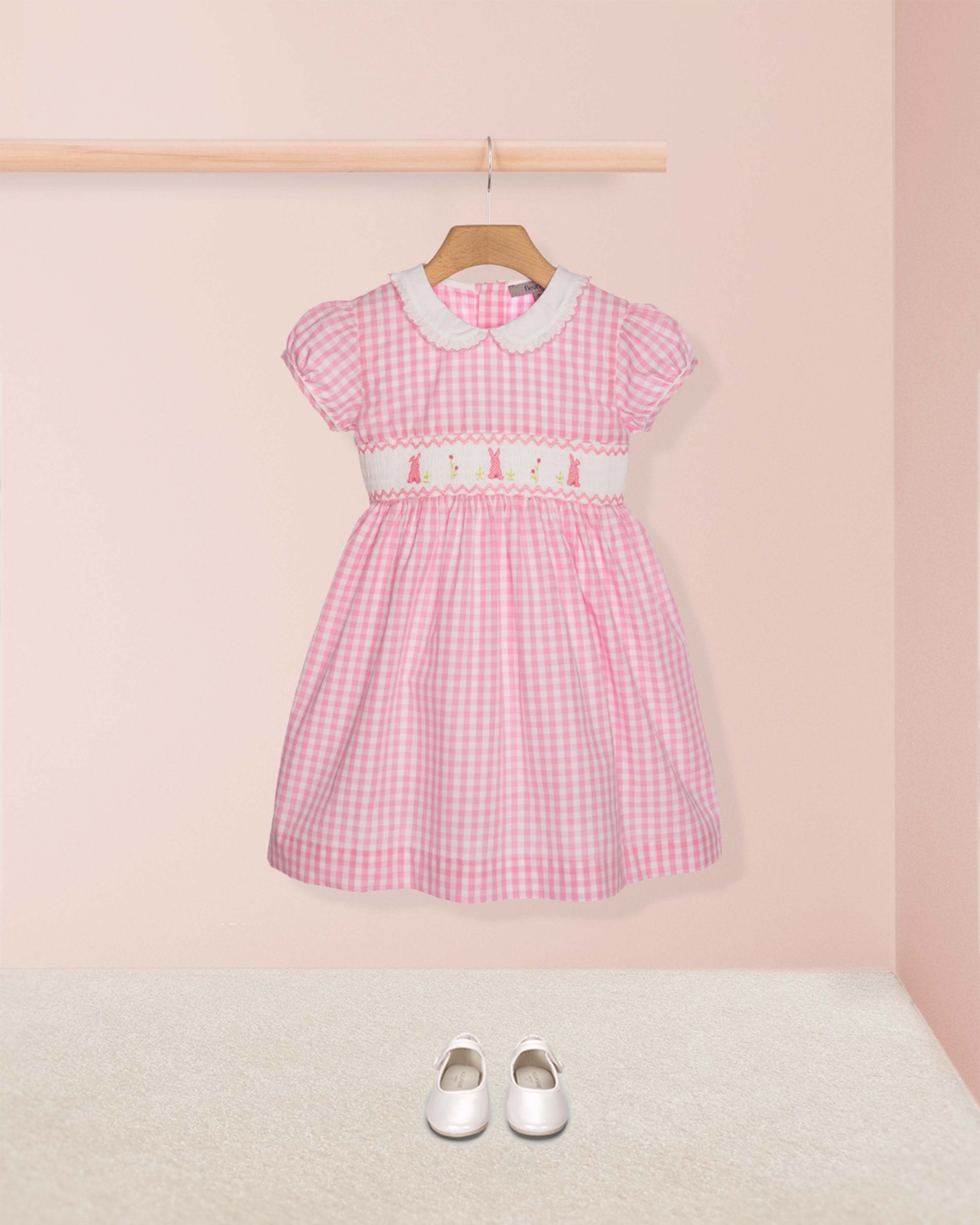 Pink checkered dress with white collar and belt on a hanger against a pink wall.